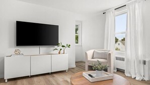 A modern living room with a flat-screen TV, a white cabinet, a round wooden coffee table, a beige sofa, and a window with white curtains. - Halcyon Haven - A Heavenly Homestead for 4 (Morton)