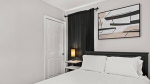 A bedroom with a bed, a nightstand, a lamp, a door, and a wall-mounted abstract art piece.