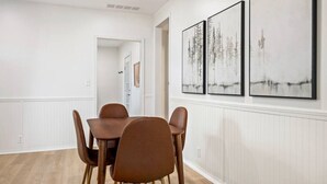 Een eetruimte met een houten tafel en bruine stoelen, witte muren en drie ingelijste kunstwerken aan de muur.