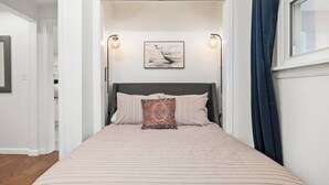 Een slaapkamer met een bed, twee wandlampen, een kunstwerk in een lijst boven het bed, en een raam met gordijnen.