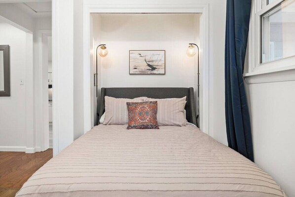 A bedroom with a bed, two wall-mounted lamps, a framed artwork above the bed, and a window with curtains.