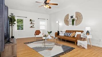 A spacious living room with a brown leather sofa, a coffee table, and a ceiling fan. There are shelves with plants and a large window providing natural light.