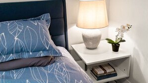 Een netjes opgemaakt bed met een blauw-witte bedspreide en kussens, een wit nachtkastje met een lamp, een kleine potorkidee en een stapel boeken.