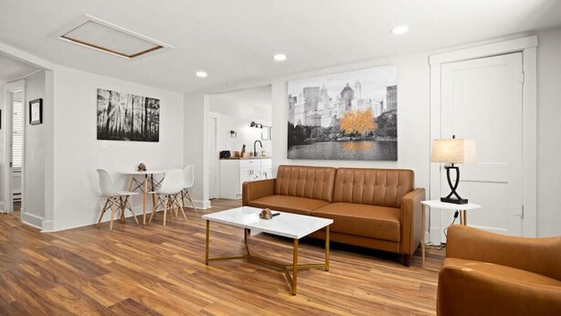 A modern living room with a brown sofa, a white coffee table, and a dining area with white chairs. There is a kitchen in the background with a sink and cabinets.