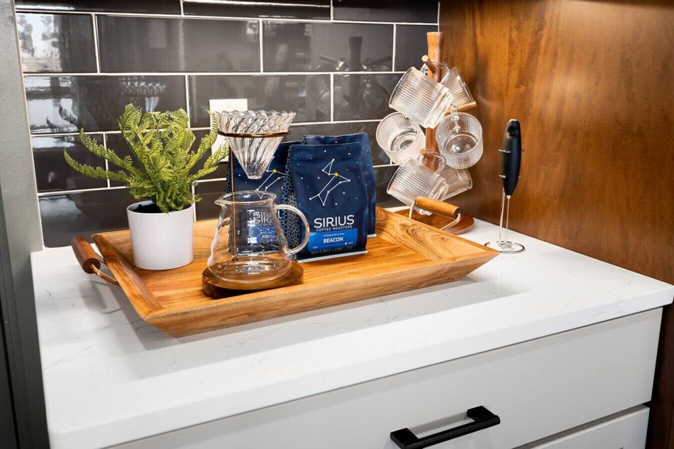 Una encimera de cocina con una cafetera, una planta en maceta, una jarra de vidrio, un paquete de café y una bandeja de madera.