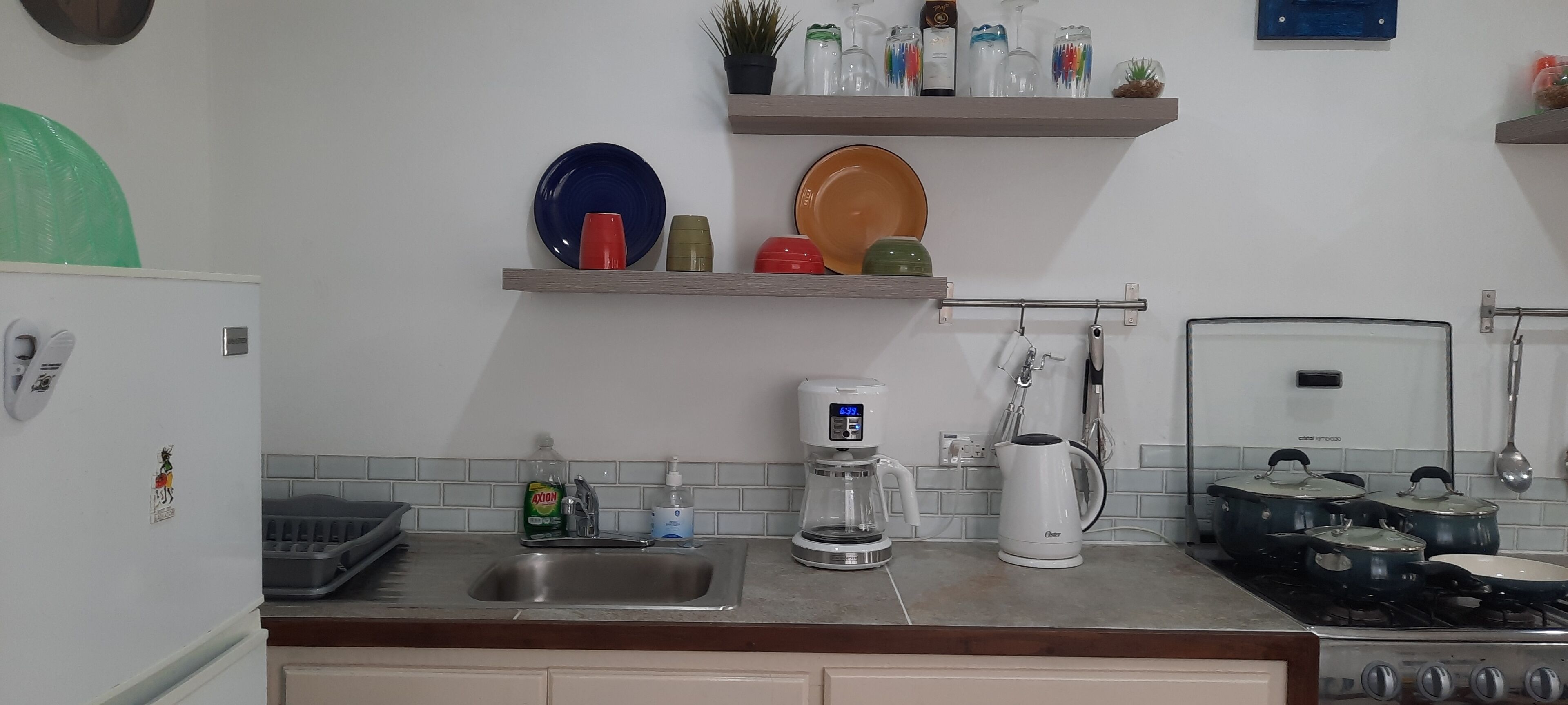 A kitchen with a sink, coffee maker, kettle, and stove. There are shelves above the counter holding plates and jars.