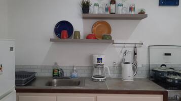 A kitchen with a sink, coffee maker, kettle, and stove. There are shelves above the counter holding plates and jars.