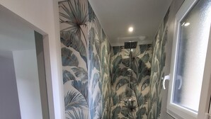 Une petite salle de bain aux murs ornés d’un motif de feuilles tropicales, avec un espace douche et une fenêtre en verre dépoli.