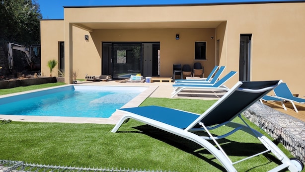 Un espace piscine moderne avec des chaises longues, une piscine à l’eau d’un bleu limpide et un bâtiment de couleur beige doté d’une porte en verre.