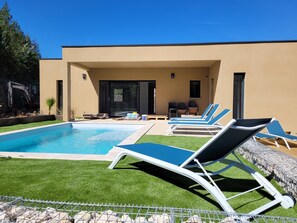 Un espace piscine moderne avec des chaises longues, une piscine à l’eau d’un bleu limpide et un bâtiment de couleur beige doté d’une porte en verre.