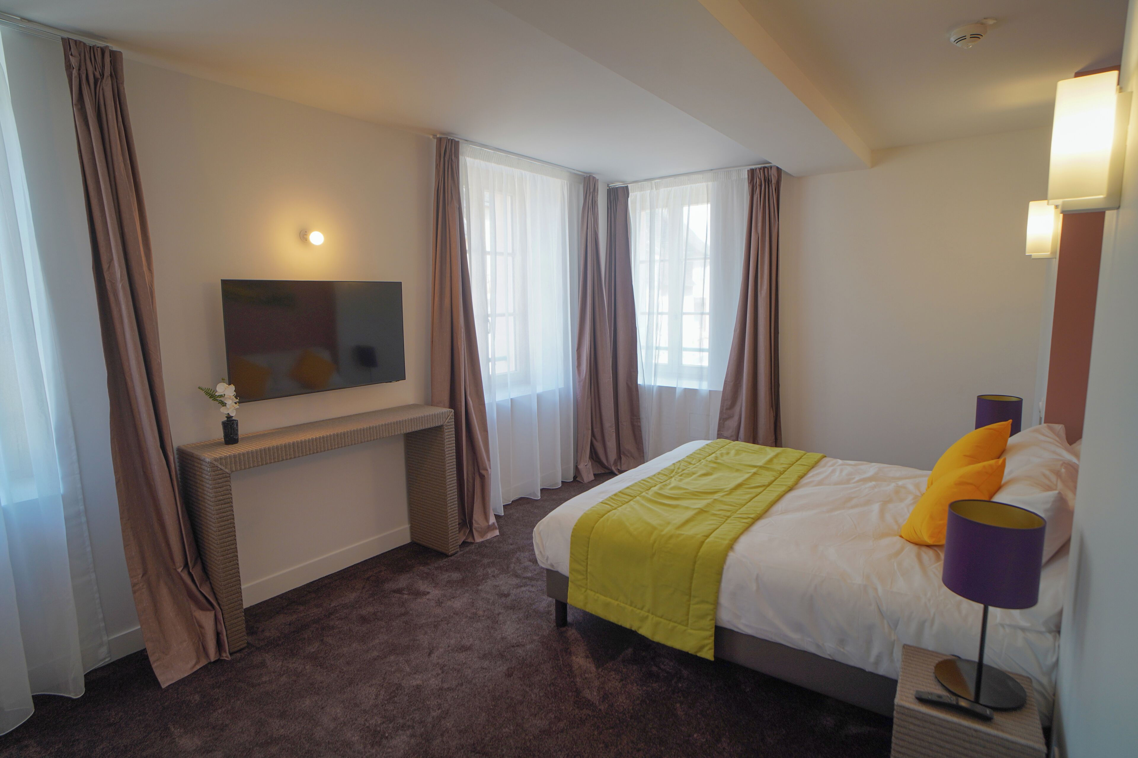 Ein Hotelzimmer mit Bett, Wandfernseher, Nachttisch mit Lampe und einem Fenster mit Vorhängen.