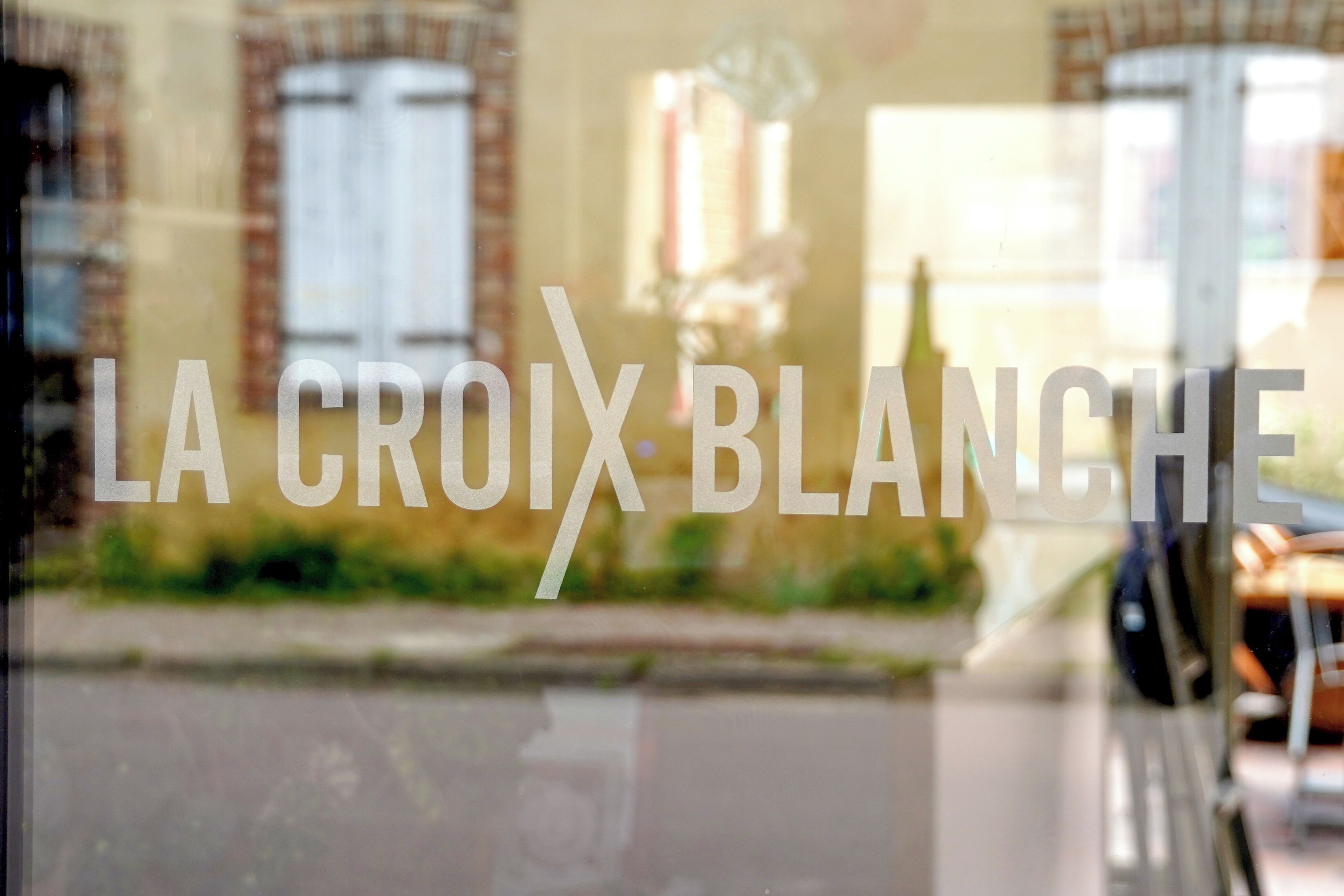 Una ventana con el texto "LA CROIX BLANCHE" y una vista borrosa de un edificio y una calle al exterior.