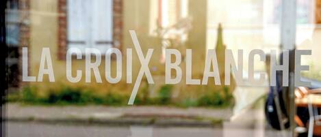Una ventana con el texto "LA CROIX BLANCHE" y una vista borrosa de un edificio y una calle al exterior.