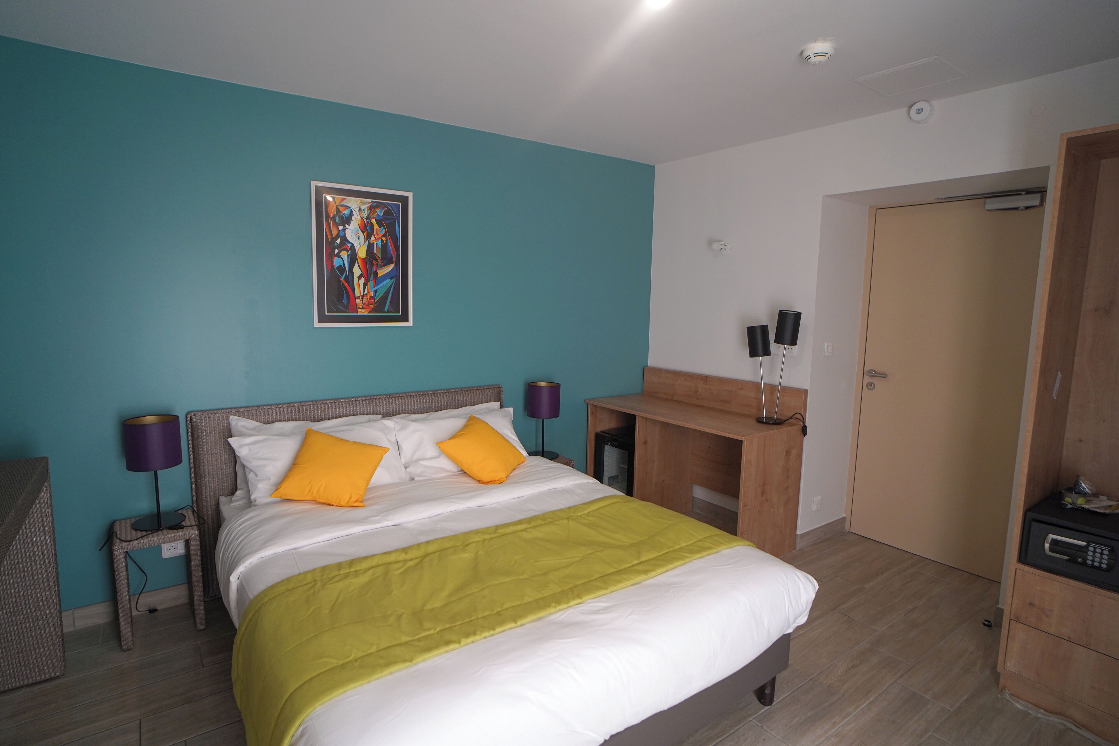 A hotel room with a bed, a desk, a chair, a wardrobe, and a painting on the wall.