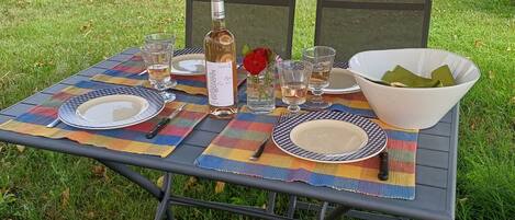 Mesa exterior con vajilla, vasos, una botella y un bol, rodeada de sillas y vista a un área verde con árboles.