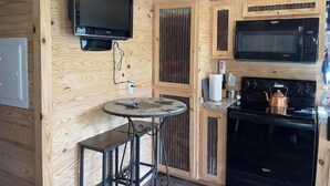 A compact kitchen with wooden walls, a black microwave, a stove, and a small round table with two stools. - Rustic Mountain Cabin 225 (Topton)