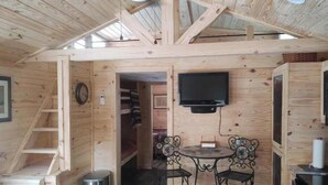 A wooden cabin interior with a loft bedroom, a dining area with two chairs, a TV mounted on the wall, and a small table. - Rustic Mountain Cabin 225 (Topton)