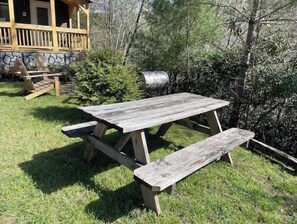 Una mesa de picnic de madera rústica con bancos, ubicada en un espacio cubierto de césped, con un muro de piedra y una terraza de madera al fondo.