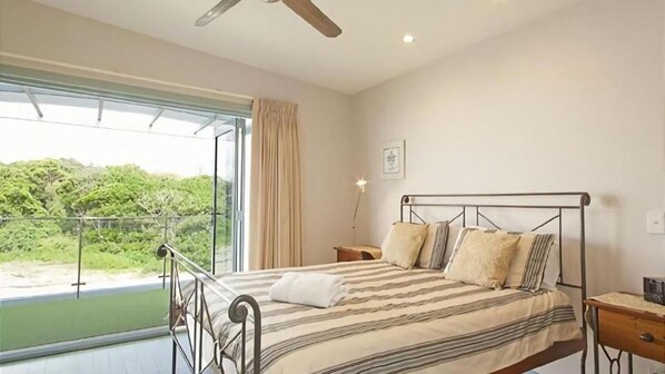 Ein Schlafzimmer mit Bett, Nachttisch, Fenster mit Blick auf Grünflächen und Deckenventilator.