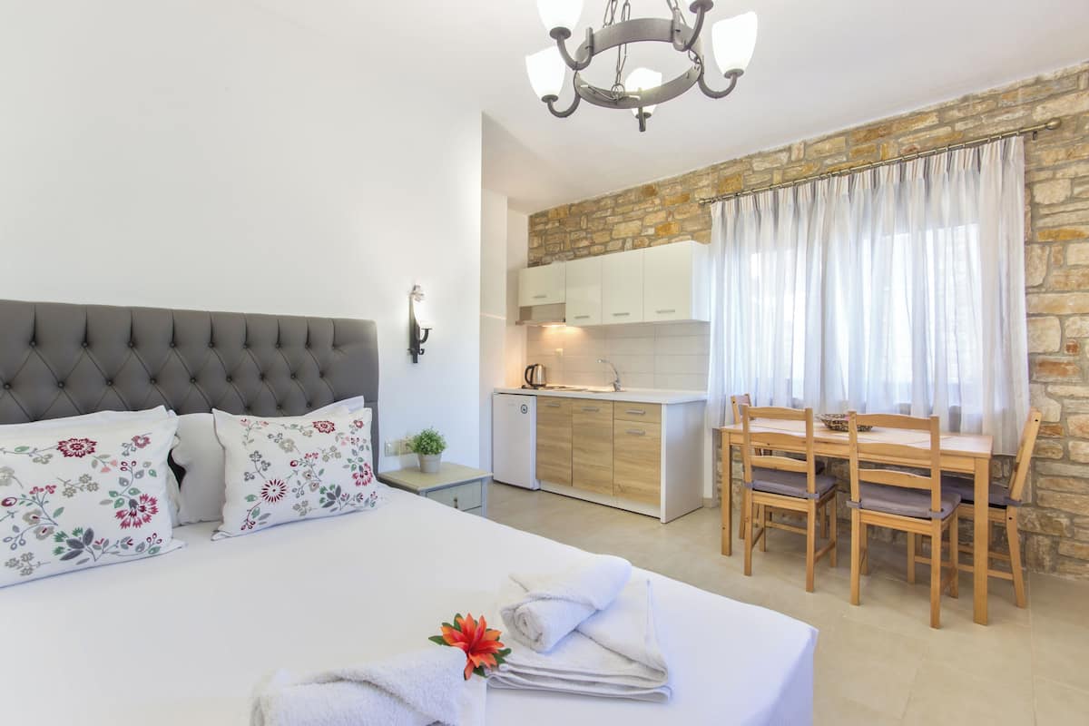 Une chambre à coucher avec une tête de lit capitonnée, des coussins à motifs floraux, un lit bien fait avec des serviettes, un coin repas avec des chaises en bois, un mur en pierre et une cuisine avec des meubles blancs et un évier.