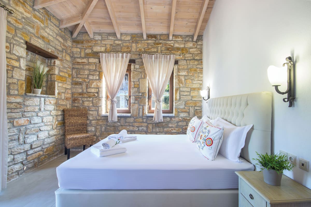 Une chambre avec un mur en pierre, un lit recouvert de draps blancs, un plafond en bois et une table de chevet agrémentée d’une plante.