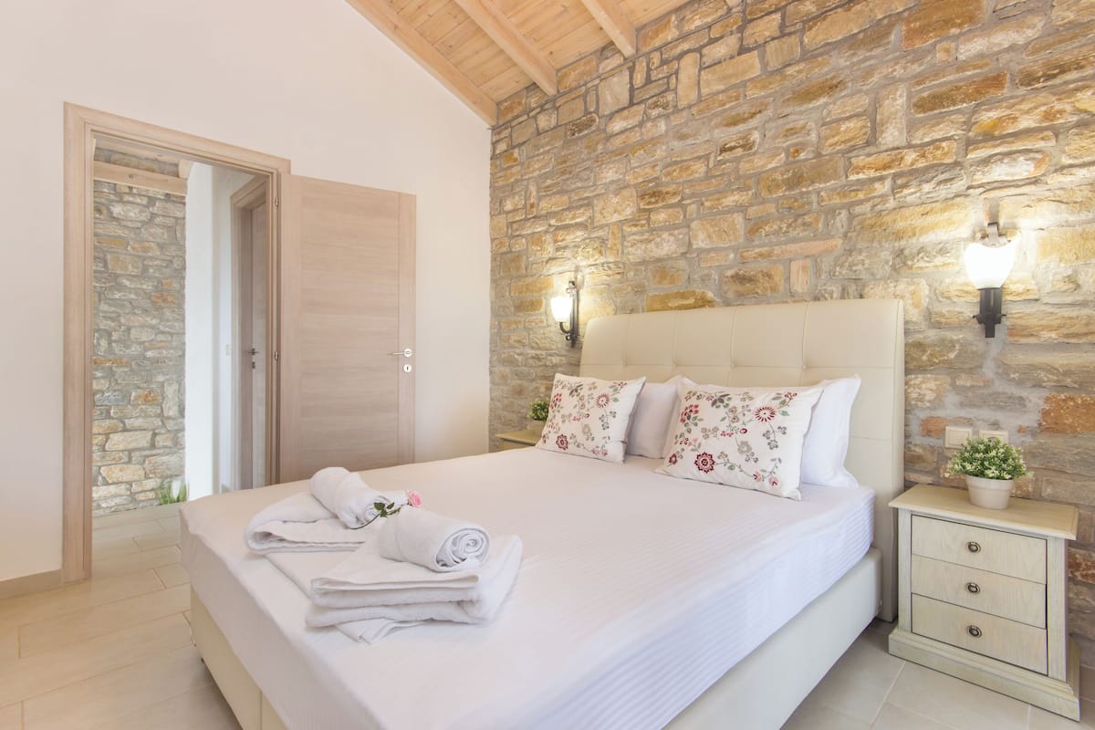 Une chambre avec un mur en pierre, un plafond en bois, un lit avec du linge de lit blanc et une table de chevet agrémentée d’une plante.