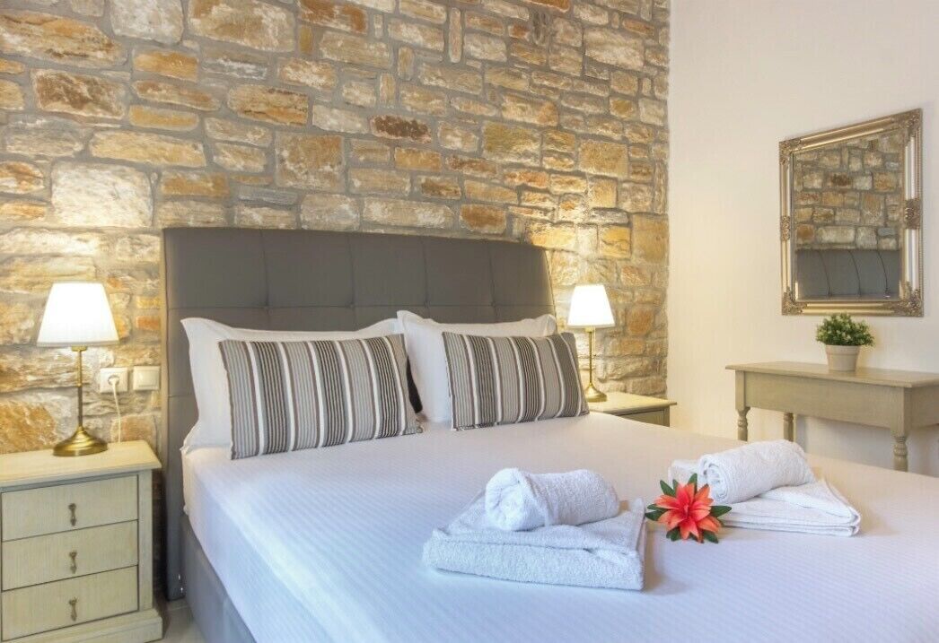 A hotel room with a stone wall, a bed with striped pillows, two bedside lamps, a nightstand, and a small table with a plant.