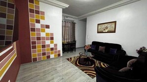 Un salon avec un canapé noir, une table basse et un tapis à motifs. On aperçoit une chaise au fond et un tableau accroché au mur.
