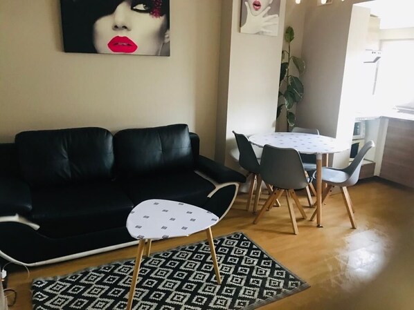 Ein Wohnzimmer mit einem schwarzen Ledersofa, einem kleinen weißen Tisch und einem Teppich mit Muster sowie ein Essbereich mit einem runden Tisch und Stühlen.