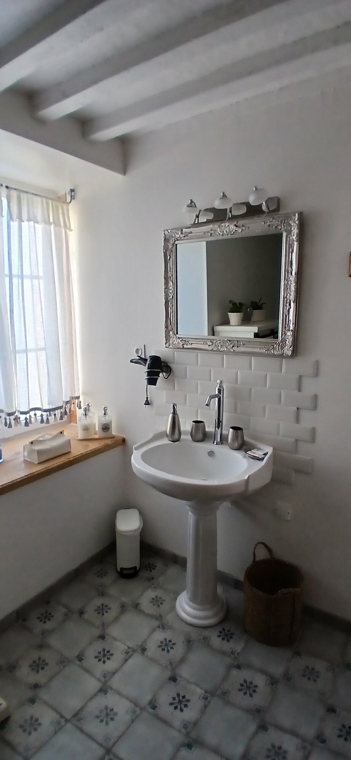 A bathroom with a white sink, a mirror with a decorative frame, a window with curtains, and a patterned floor.
