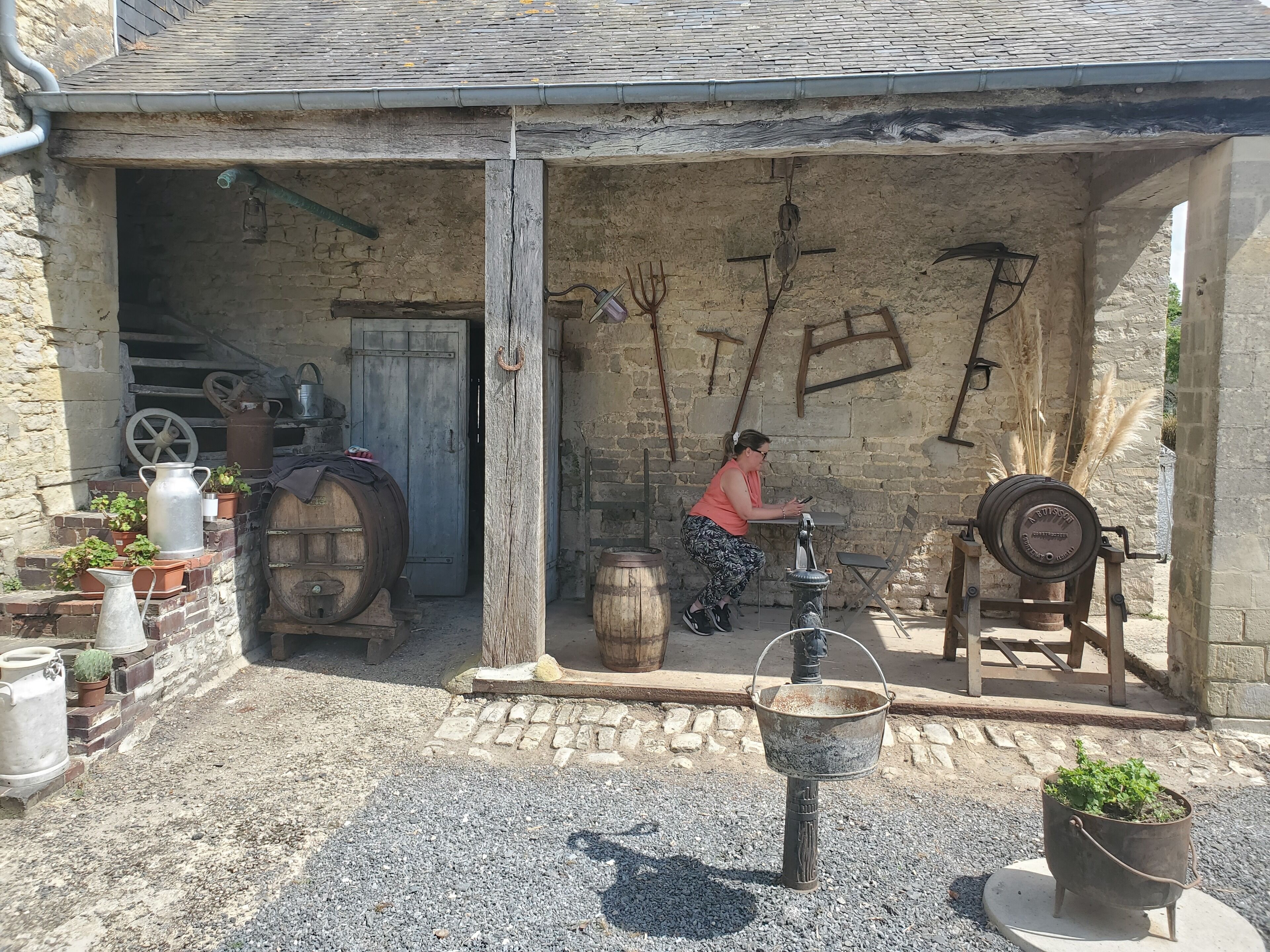 Un edificio rústico de piedra con una puerta de madera, un barril metálico y una rueda.