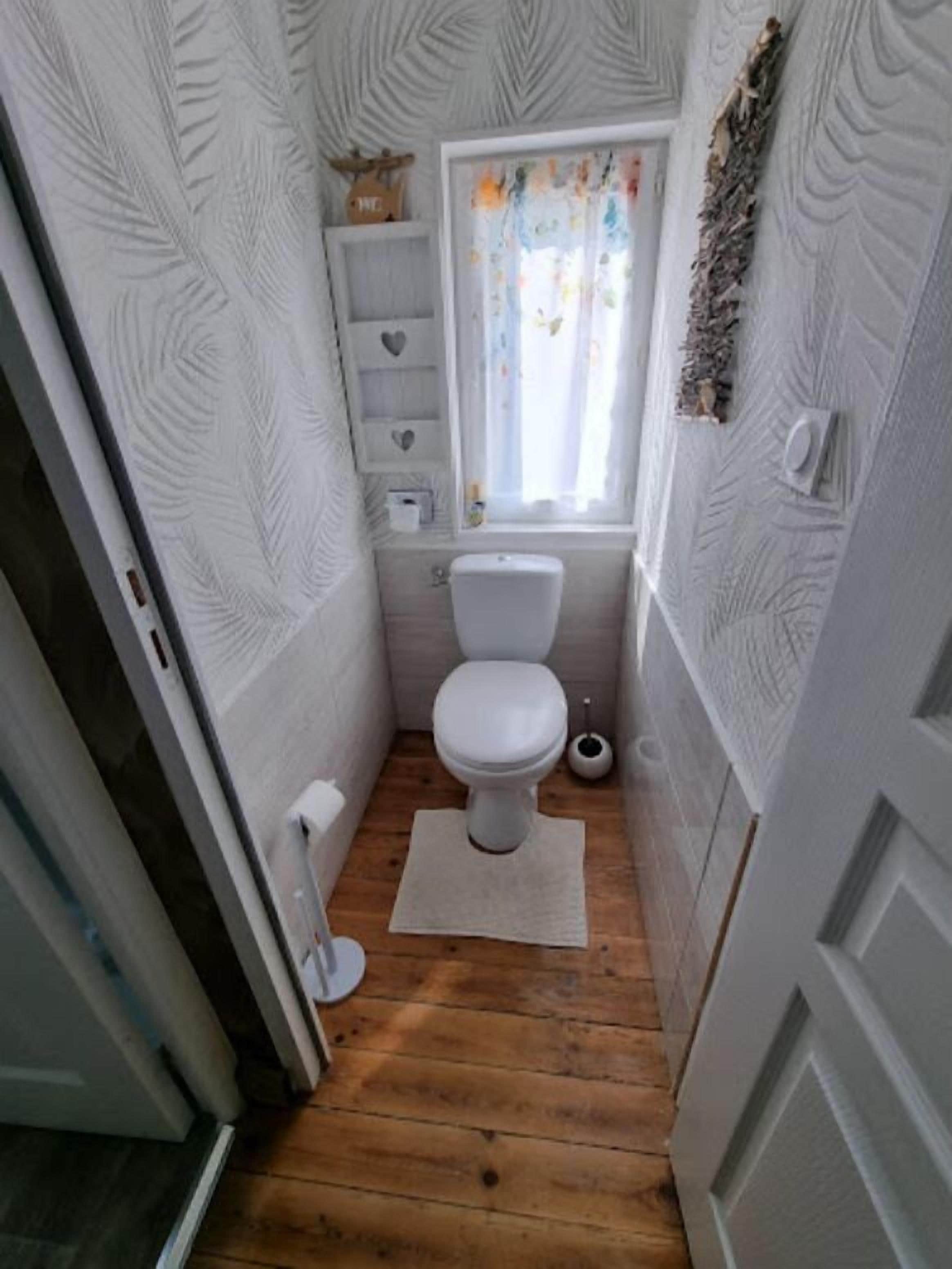 A small, compact bathroom with a white toilet, a wooden floor, and a window with a curtain.