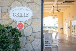 Un muro de piedra con un letrero que indica Cinnamon Beach Grill, mesas al aire libre y una vista despejada del entorno.