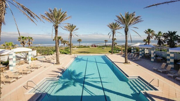 Zona de piscina con sillones de descanso y palmeras, con vista a una playa y el océano.