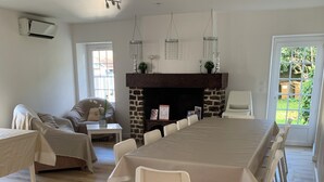 A spacious dining area with a long table and white chairs, a stone fireplace, and a view of greenery outside.