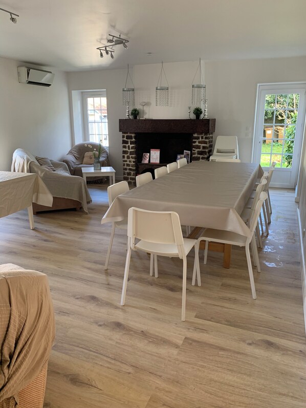 A spacious dining area with a long table and white chairs, a stone fireplace, and a view of greenery outside.