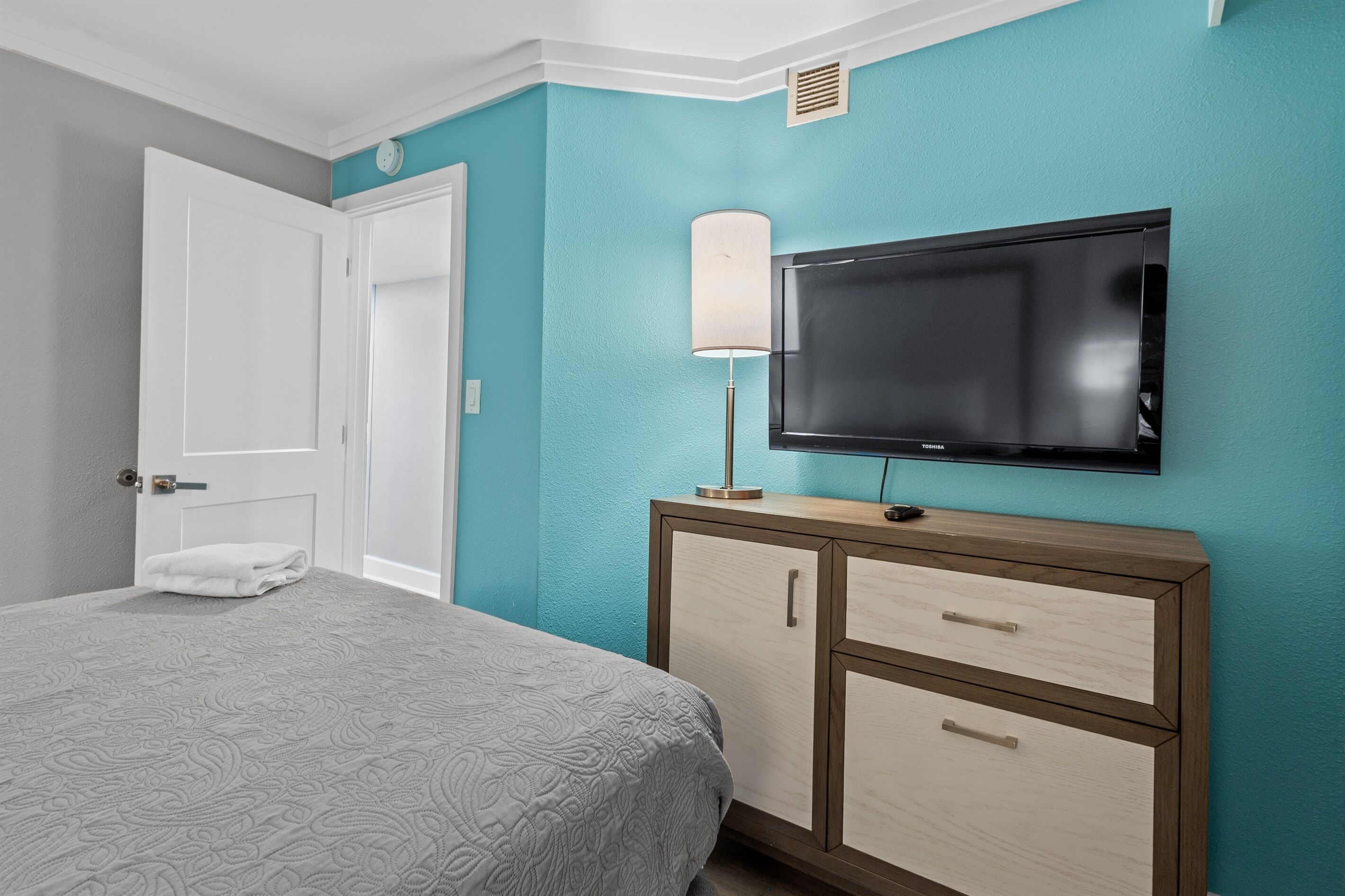 Quarto de hotel com cama, televisão na parede, mesa de cabeceira e abajur, além de uma porta.