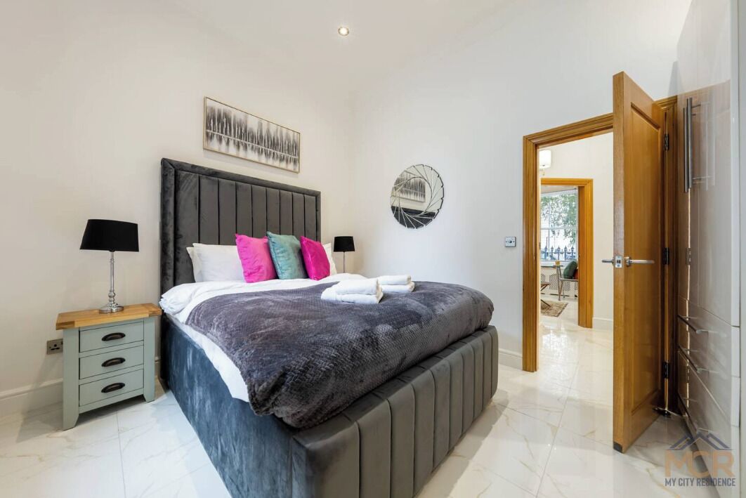 A bedroom with a large bed, a bedside table, a wall-mounted mirror, and a door leading to another room.