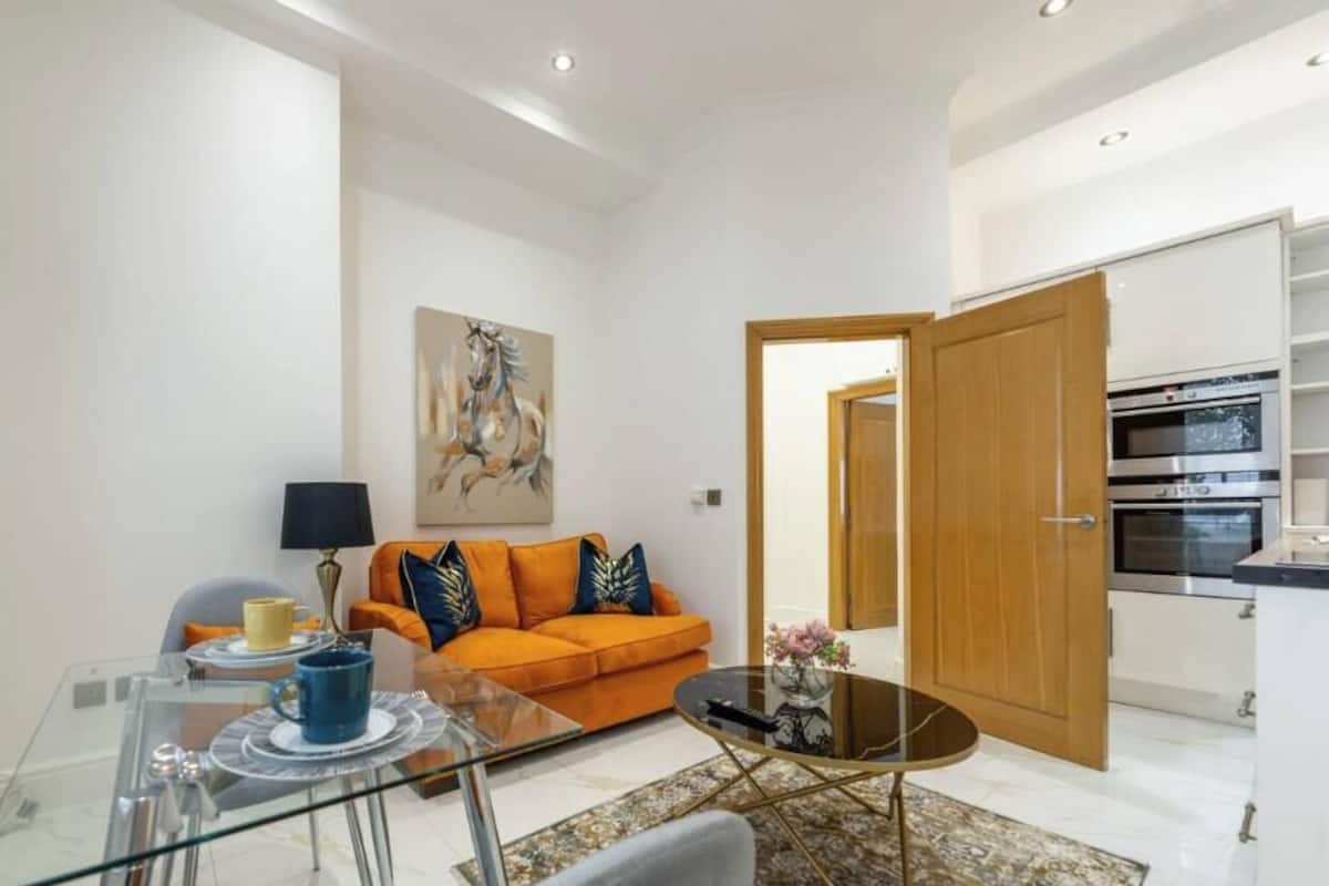 Un salon moderne avec une table basse en verre, un canapé agrémenté de coussins orange, un tableau accroché au mur et un coin cuisine visible à travers une porte ouverte.