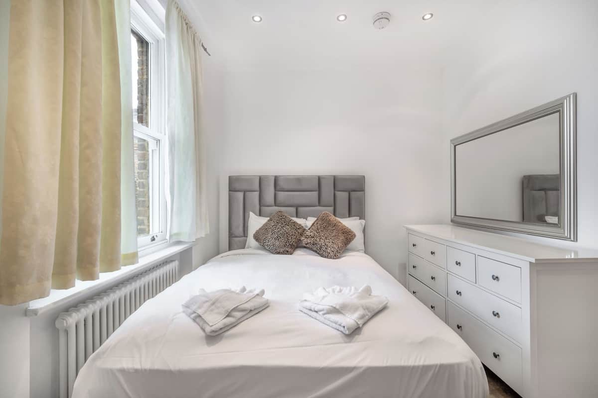 Une chambre bien rangée, avec un grand lit, une commode, un miroir et une fenêtre avec des rideaux.