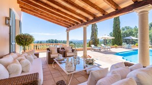 Un espace extérieur aménagé, abrité et doté de mobilier en osier, d’une table en verre, et offrant une vue sur une piscine et un paysage verdoyant.