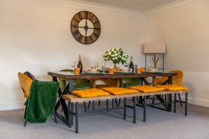 A dining area with a wooden table, green and yellow chairs, a bench with yellow cushions, a wall clock, and a vase with flowers. - City Chic: 3 Bedroom, 3 Bathroom Luxury Living (Greater London)