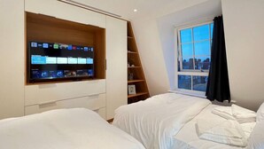 Ein modernes Hotelzimmer mit einem großen Flachbildfernseher an einer Holzablage, zwei Betten mit weißer Bettwäsche, einem Fenster mit Blick auf die Stadt und einer kleinen Ablage mit persönlichen Gegenständen.