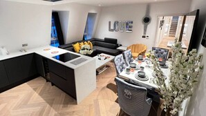 Eine moderne Küche mit einer zentralen Kochinsel, ein Essbereich mit Tisch für vier Personen und eine Wohnbereits mit einer Couch und einem dekorativen Wandbild.