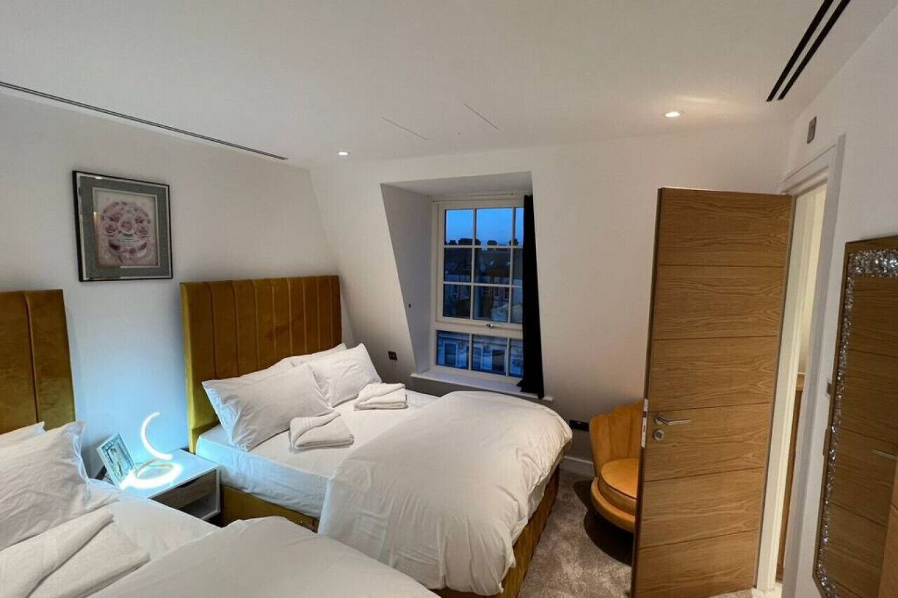 A bedroom with two beds, a bedside table with a lamp, a window with a view of buildings, and a wooden door.