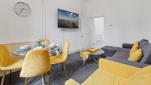 Un salon moderne avec un coin repas, comprenant une table ronde, des chaises jaunes, un canapé gris et un tableau représentant un paysage urbain accroché au mur.