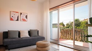 Ein modernes Wohnzimmer mit einem grauen Sofa, zwei Kissen, einem gewebten Hocker und einer großen Schiebeglasdoor, die auf einen Balkon mit Blick auf Grünflächen und Gebäude führt.