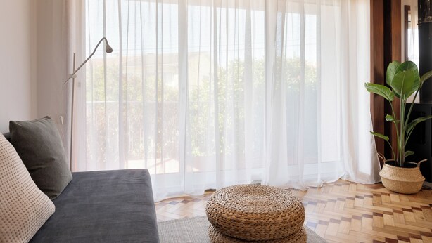 Ein Zimmer mit grauem Sofa, einem gewebten Hocker, einer Zimmerpflanze und transparenten Vorhängen.
