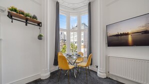 Un coin repas avec une table ronde et quatre chaises, une fenêtre avec des rideaux, un téléviseur fixé au mur et une étagère avec des plantes.