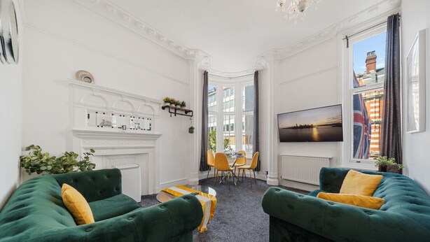 Ein Zimmer mit zwei grünen Sofas, einem Kamin, einem Essbereich mit gelben Stühlen und einem an der Wand befestigten Fernseher.